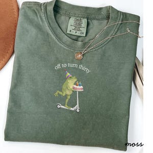 May include: Green Comfort Colors t-shirt with the text "off to turn thirty" and a graphic of a frog riding a scooter and carrying a birthday cake. A gold necklace is draped across the shirt.