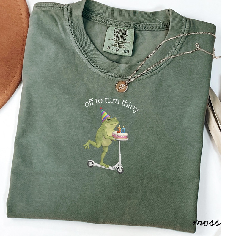 May include: Green Comfort Colors t-shirt with the text "off to turn thirty" and a graphic of a frog riding a scooter and carrying a birthday cake. A gold necklace is draped across the shirt.