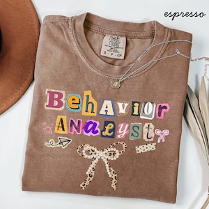 Könnte beinhalten: Espressofarbenes T-Shirt mit den Worten "Behavior Analyst" in einem farbenfrohen, ausgeschnittenen Stil. Das Shirt hat eine Schleife mit Leopardenmuster und eine silberne Halskette.