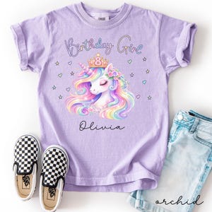 Pode incluir: Uma camiseta lilás com um gráfico de unicórnio colorido, uma coroa e o texto "Birthday Girl" e "Olivia". A imagem inclui tênis xadrez e shorts jeans. Roupa infantil.