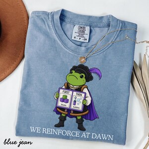 Op de afbeelding: Een blauw t-shirt met ronde hals en een afbeelding van een groene kikker in historische kleding met een paarse cape en hoed. De kikker houdt een bord vast met FIRST THEN en illustraties van blokken en bellen. De tekst WE REINFORCE AT DAWN staat eronder gedrukt.