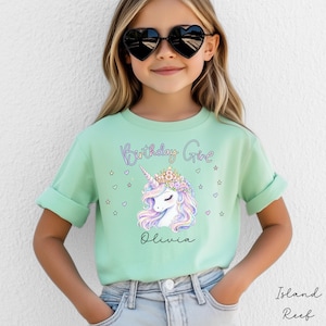 Peut inclure: T-shirt vert menthe avec un motif de licorne, une couronne de fleurs et le texte "Birthday Girl" au-dessus et "Olivia" en dessous. Le t-shirt a des manches retroussées. La personne porte des lunettes de soleil en forme de cœur.
