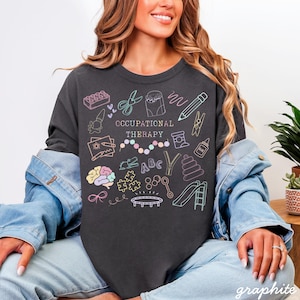 Pu&ograve; includere: T-shirt color grafite con un'illustrazione colorata di strumenti di terapia occupazionale e la scritta "OCCUPATIONAL THERAPY". Il design include un cervello, forbici, mattoncini e una matita.