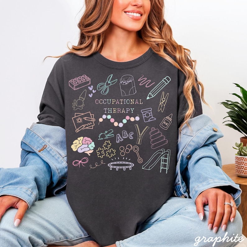 Pu&ograve; includere: T-shirt color grafite con un'illustrazione colorata di strumenti di terapia occupazionale e la scritta "OCCUPATIONAL THERAPY". Il design include un cervello, forbici, mattoncini e una matita.