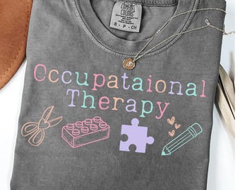 Puede incluir: Camiseta gris con el texto "Occupational Therapy" en colores arcoíris. La camiseta presenta un par de tijeras, una pieza de rompecabezas, un bloque de bloques de construcción y un lápiz con corazones.