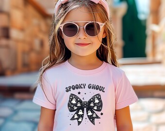 Puede incluir: Una camiseta de manga corta de color rosa claro con un gráfico de lazo negro con patrones de fantasmas y estrellas blancos. El texto "SPOOKY GHOUL" está impreso en negro sobre el lazo. La camiseta se lleva con una diadema rosa claro a juego.