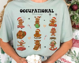May include: A light blue t-shirt with the words "OCCUPATIONAL therapy" above gingerbread people in Santa hats. The gingerbread people are engaged in various activities, including skateboarding, swinging, and balancing on a ball.