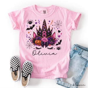 Peut inclure: Un t-shirt rose clair avec un motif de licorne portant une couronne noire, une citrouille, des fleurs, des chauves-souris, des araignées et des toiles d'araignée. Le chiffre 8 est centré dans la couronne et le prénom Olivia est écrit en noir.