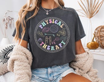 Puede incluir: Camiseta gris oscuro con un diseño circular con las palabras "PHYSICAL THERAPY" en colores pastel. El diseño incluye ilustraciones de equipos de ejercicio. La camiseta es de un estilo cómodo e informal.