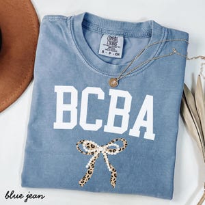 Könnte beinhalten: Ein blaues T-Shirt in Jeansoptik mit Rundhalsausschnitt und einem Comfort Colors Markenetikett. Auf der Vorderseite befinden sich die weißen Blockbuchstaben BCBA über einer Grafik einer Leopardenmuster-Schleife mit weißen Perlen. Eine Goldkette liegt darauf.