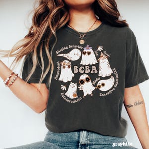 Op de afbeelding: Donkergrijs t-shirt met een cirkelvormig ontwerp met spookillustraties en de tekst "BCBA". Het ontwerp bevat woorden als "Antecedent", "Reinforcement" en "Executive Functioning". Het shirt is gemaakt van zacht materiaal.