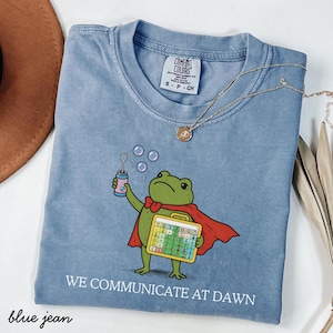May include: A blue jean coloured t-shirt featuring a green frog wearing a red cape, holding a bubble wand and a communication board. The text "WE COMMUNICATE AT DAWN" is printed below the graphic. The shirt has a Comfort Colors brand label inside.