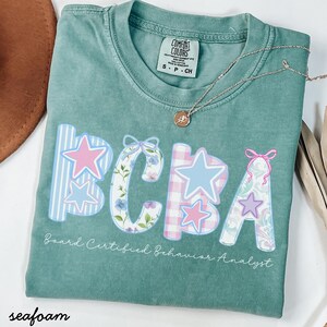 Op de afbeelding: Zeegroen t-shirt met de letters "BCBA" in een decoratief lettertype, waarbij elke letter een ander patroon en kleur heeft, waaronder sterren en bloemmotieven. De tekst "Board Certified Behavior Analyst" staat onder de letters. Een zilveren ketting met een hanger ligt op het shirt.