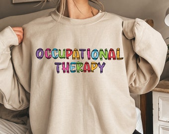 Puede incluir: Una sudadera beige con el texto "OCCUPATIONAL THERAPY" impreso en letras de colores. Las letras están delineadas en negro y tienen un patrón de arcoíris.