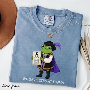 Könnte beinhalten: Ein blaues T-Shirt mit einer Grafik eines grünen Frosches in historischer Kleidung, der eine Brille trägt und eine Sehtafel hält. Der Frosch trägt einen schwarzen Hut mit violetter Feder, eine schwarze Tunika mit Goldbesatz und einen violetten Umhang. Text: WE SAVE EYES AT DAWN.