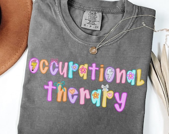 May include: A gray Comfort Colors t-shirt with the words "Occupational Therapy" in colorful, playful lettering. The design includes lightning bolts, flowers, and bows. A silver necklace with a pendant is draped across the shirt.
