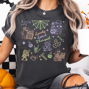 May include: Dark gray t-shirt with a colorful Halloween-themed graphic. The design includes a ghost, cat in a witch hat, spiderweb, and the text "Spooky Speech Therapist". Other illustrations include a mummy, skeleton, and puzzle pieces.