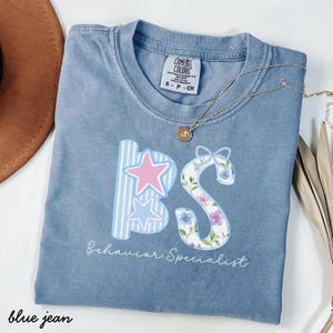 Op de afbeelding: Een blauwe Comfort Colors t-shirt met de letters "BS" in een decoratief ontwerp. De "B" heeft blauw-witte strepen en roze en blauwe sterren. De "S" is versierd met bloemen. De woorden "Behavior Specialist" staan eronder.