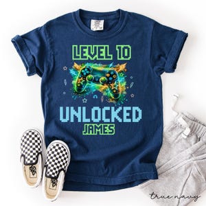 May include: Navy blue t-shirt with a video game controller graphic. The shirt reads "LEVEL 10 UNLOCKED JAMES" in pixelated font. A pair of black and white checkered slip-on sneakers and gray sweatpants are also in the image.