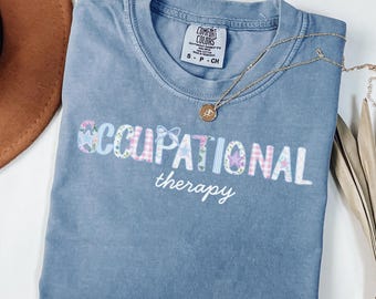 Puede incluir: Una camiseta azul vaquera con las palabras "OCCUPATIONAL therapy" en una fuente decorativa. La palabra "OCCUPATIONAL" está en un diseño de patchwork con patrones florales y de estrellas. Un collar dorado con un colgante está drapeado sobre la camisa.