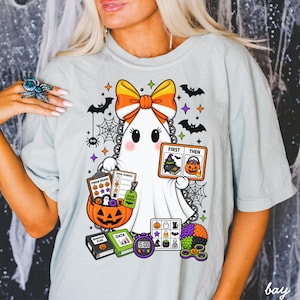 Könnte beinhalten: Ein hellgraues T-Shirt mit einer Grafik eines weißen Geistes, der eine Schleife im Candy-Corn-Design trägt. Der Geist hält ein Planungsboard und ein Token-Board. Ergänzt wird das Motiv durch einen Kürbis, Fledermäuse, Spinnennetze und Bücher.