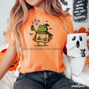 Könnte beinhalten: Ein orangefarbenes T-Shirt mit einer Grafik eines grünen Frosches, der einen Hexenhut und einen Umhang trägt und einen Seifenblasenstab hält. Der Frosch hält ein Schild mit der Aufschrift FIRST THEN, Blöcken und Süßigkeiten. Darunter steht WE REINFORCE AT DAWN.