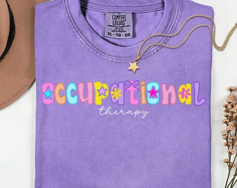 May include: A violet short-sleeved t-shirt with a crew neckline. The front features the text "occupational therapy" in a colorful, wavy, multi-font design with star and flower accents. A gold-toned necklace with a star pendant rests on the shirt.