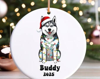 May include: White ceramic Christmas ornament featuring a Siberian Husky wearing a Santa hat and wrapped in colorful string lights. The ornament has the text "Buddy 2025" in black, and is hanging from a gold string.