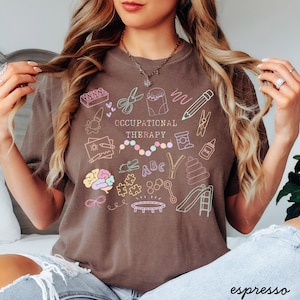 Pu&ograve; includere: T-shirt color espresso con la scritta "OCCUPATIONAL THERAPY" e illustrazioni di scarabocchi colorati. Gli scarabocchi includono un cervello, forbici, una matita e uno scivolo. La maglietta &egrave; uno stile comodo e casual.
