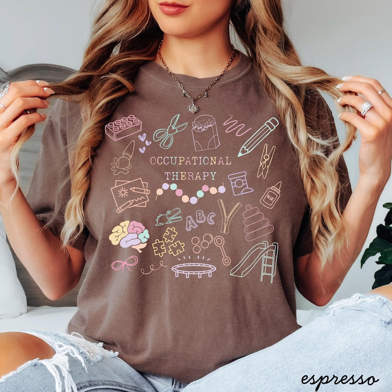 Pu&ograve; includere: T-shirt color espresso con la scritta "OCCUPATIONAL THERAPY" e illustrazioni di scarabocchi colorati. Gli scarabocchi includono un cervello, forbici, una matita e uno scivolo. La maglietta &egrave; uno stile comodo e casual.