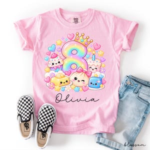 Könnte beinhalten: Ein hellrosa T-Shirt mit einer großen regenbogenfarbenen Acht, die eine goldene Krone trägt, umgeben von Cartoon-Cupcakes, einem Kuchen mit Kerze und Süßigkeiten. Der Name Olivia ist darunter in einer schwarzen Schreibschrift aufgedruckt.
