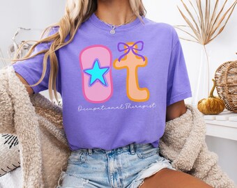 May include: A violet-colored t-shirt with the letters "OT" in a playful design. The "O" is pink with a blue star, and the "T" is orange with a purple bow. The words "Occupational Therapist" are written below the letters.