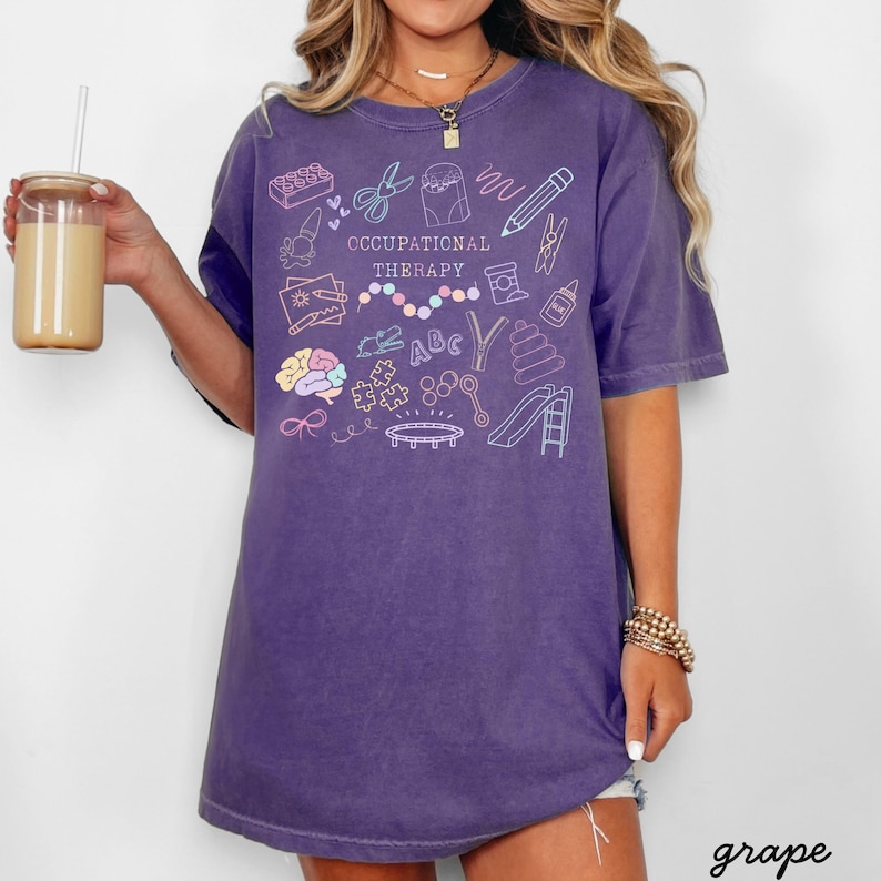 Pu&ograve; includere: Una maglietta viola con la scritta "OCCUPATIONAL THERAPY" e illustrazioni colorate di strumenti terapeutici. La maglietta &egrave; di taglio ampio e ha maniche corte. La parola "grape" &egrave; scritta in basso a destra.