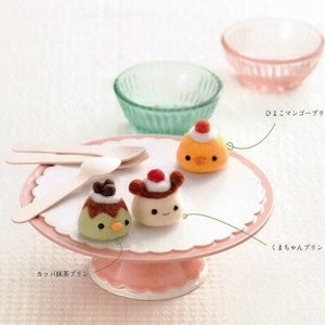 Pode incluir: Três alimentos de feltro em um suporte de bolo rosa com borda festonada. Os alimentos são um pudim verde com molho de chocolate, um pintinho amarelo com uma cereja vermelha no topo e um pudim marrom com uma cereja vermelha e um rosto branco. O suporte está sobre uma superfície branca com duas colheres de madeira.