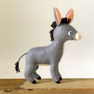 Puede incluir: Un peluche de burro gris hecho a mano con un hocico blanco, mejillas rosadas y orejas grandes y erguidas con el interior color melocotón. El burro tiene una melena y cola marrón oscuro y pezuñas negras. Está de pie sobre una superficie de madera.