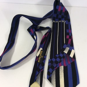 Puede incluir: Una corbata de patrón negro y azul con acentos blancos y morados. La corbata tiene un diseño geométrico y el nombre de la marca "Newman Marcus" está impreso en una etiqueta dorada.