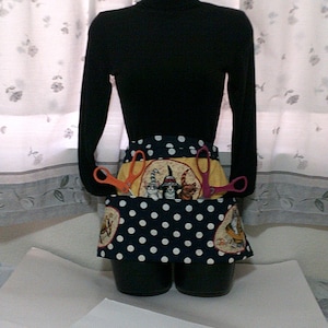 May include: A black turtleneck shirt with a white and black polka dot apron featuring a cat design. The apron has two pairs of scissors in the pockets.