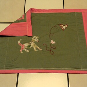 May include: A green and pink quilted throw blanket with a dog, butterfly, and heart design. The blanket has a pink border.