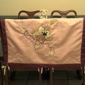 May include: A pink fabric chair cover with a brown embroidered dog design. The dog is running and the text "Dog-on-I-T!" is embroidered below it.