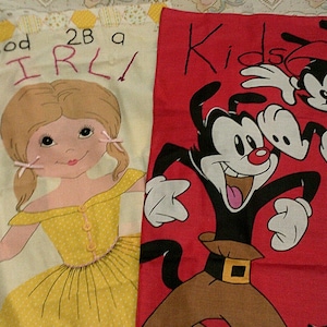 May include: A patchwork textile featuring two distinct designs. One side displays a cartoon girl in a yellow dress with the text "good 2B a GIRL!". The other side showcases cartoon characters on a red background with the text "Kids Ha".