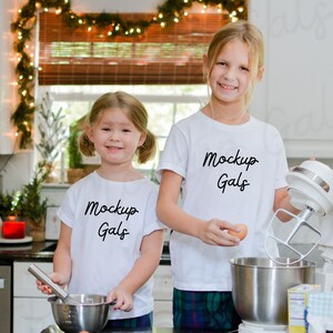 May include: Two young girls in a kitchen, wearing white t-shirts with "Mockup Gals" printed on them. One girl holds a whisk and bowl, the other holds an egg near a stand mixer. The kitchen has white cabinets and a window with a bamboo blind.