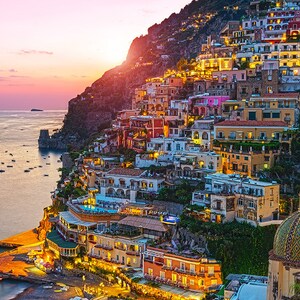 May include: A colorful town built on a cliffside overlooking a calm ocean at sunset. The town is lit up with lights, and there are boats in the water.