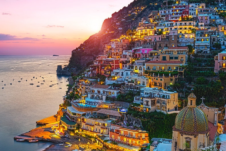May include: A colorful town built on a cliffside overlooking a calm ocean at sunset. The town is lit up with lights, and there are boats in the water.