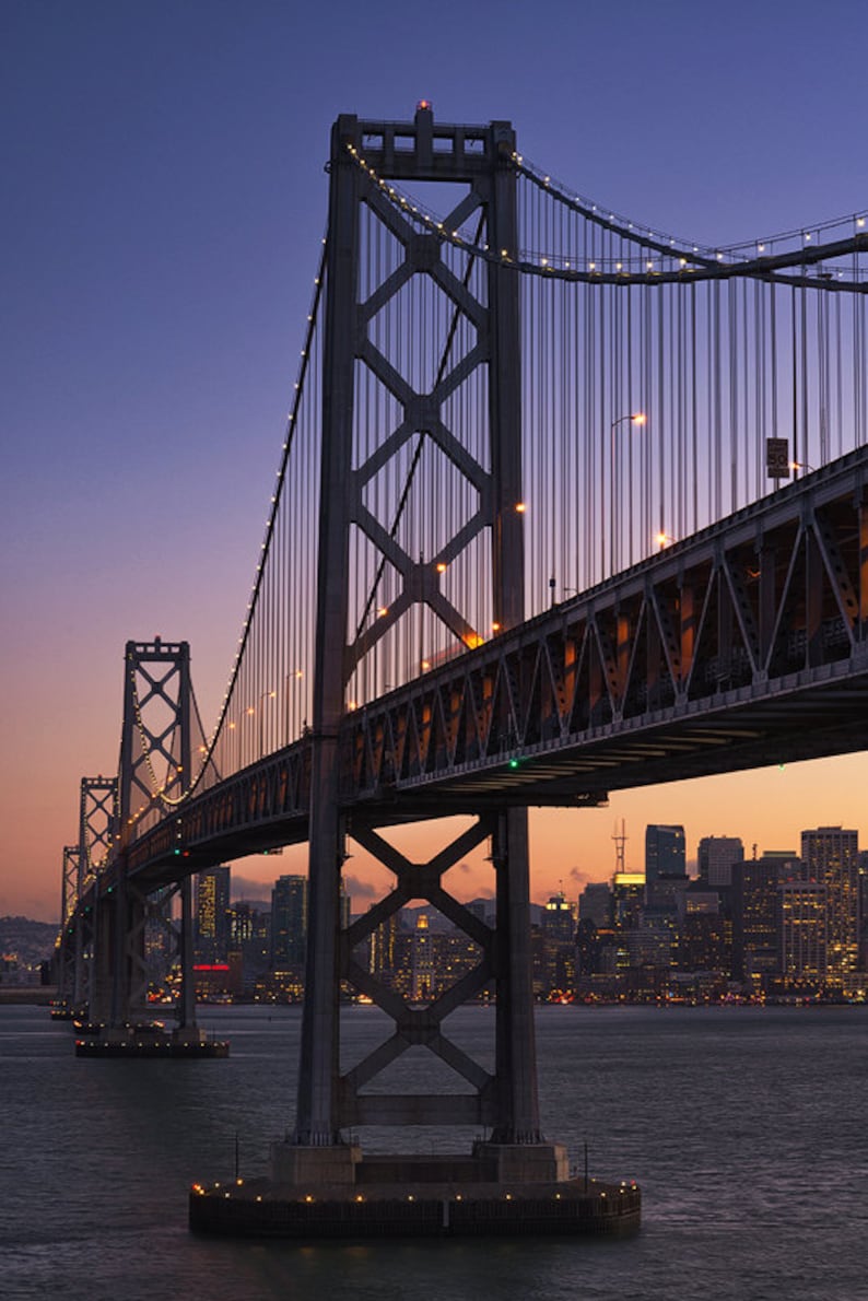 May include: A suspension bridge with a cityscape in the background. The bridge is made of steel and has a gray color. The cityscape is lit up with lights and has a purple hue. The bridge is over a body of water.