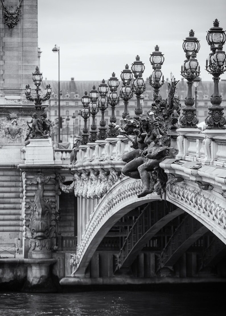 May include: A black and white photo of a bridge with ornate details and lampposts. The bridge is arched and has a stone railing with statues of figures. The bridge is over a body of water.