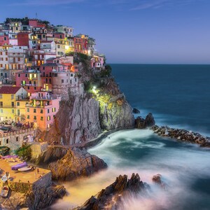 May include: A colorful village built on a cliff overlooking the ocean at dusk. The village is lit up with lights, and the water is calm and still.