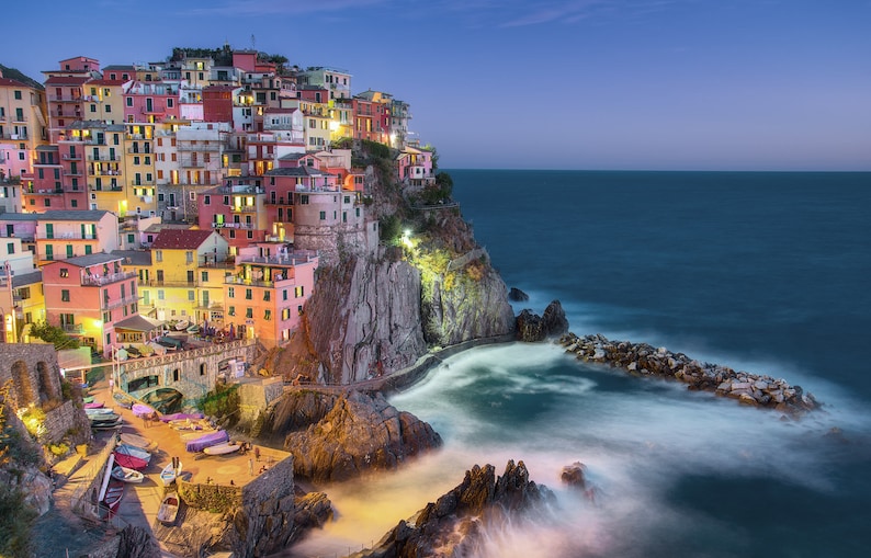 May include: A colorful village built on a cliff overlooking the ocean at dusk. The village is lit up with lights, and the water is calm and still.