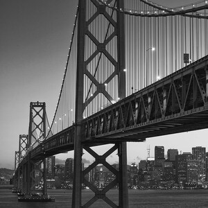 May include: A black and white photo of the Bay Bridge in San Francisco, California. The bridge is a suspension bridge with a large tower in the center. The bridge is lit up at night, and the city skyline is visible in the background.