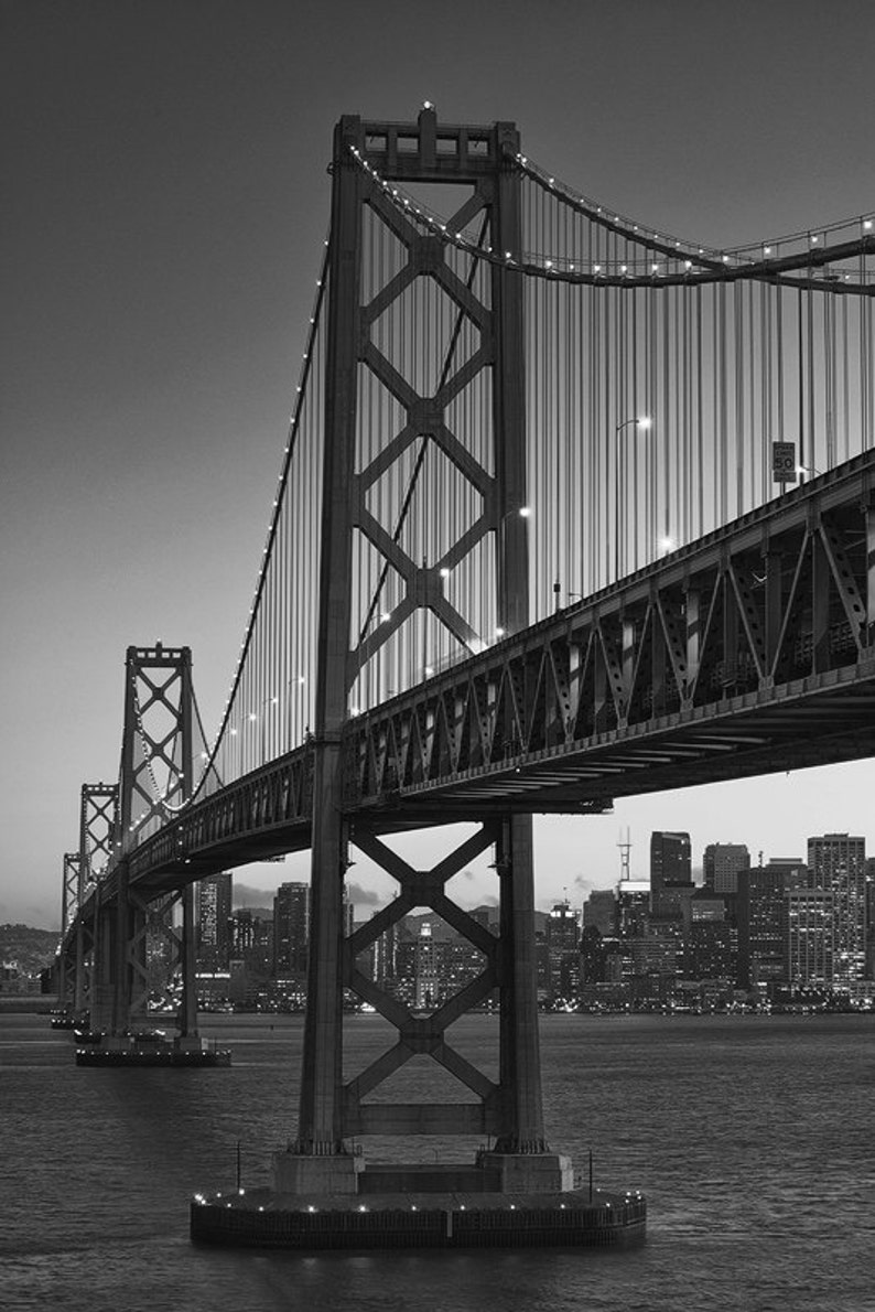 May include: A black and white photo of the Bay Bridge in San Francisco, California. The bridge is a suspension bridge with a large tower in the center. The bridge is lit up at night, and the city skyline is visible in the background.
