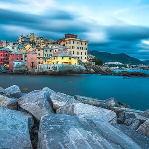 May include: A colorful Italian village with pastel-colored buildings sits on a rocky coastline. The water is a deep blue and the sky is a light blue with white clouds. The village is surrounded by lush green hills.